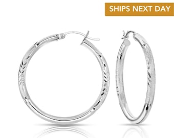 May include: A pair of silver hoop earrings featuring a textured, frosted finish with subtle, repeating etched leaf-like patterns along the outer surface. The earrings have a hinged post closure mechanism and a polished, reflective inner edge.