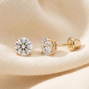May include: A pair of gold stud earrings featuring round, brilliant-cut clear stones. One earring is shown from the front, displaying the faceted stone, while the other is shown from the side, revealing the gold setting, threaded post, and screw back.