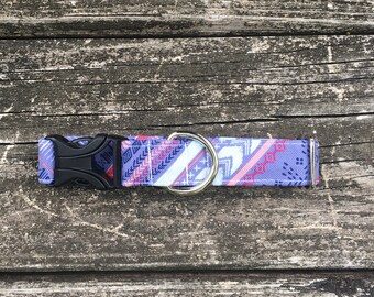 May include: A patterned dog collar with a black plastic buckle and a silver metal D-ring. The collar features a geometric design in shades of purple, pink, and blue. The collar is laying on a weathered wooden surface.