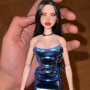 Puede incluir: Una muñeca de moda con pelo largo negro, ojos azules y labios rojos. La muñeca lleva un minivestido azul brillante y un collar dorado. La muñeca tiene la piel clara y pecas. La muñeca es un objeto de colección.