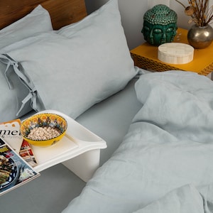 May include: A bed with light blue linen bedding, including pillows with tie closures. A white tray holds a bowl of food and magazines, including "Gourmet" and "Cucina Italiana". A yellow side table has a green Buddha head and decorative items.