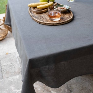 May include: A gray linen tablecloth with a woven tray holding a glass of amber liquid, green apples, and bananas. The tray is on a wooden table with a stone floor.