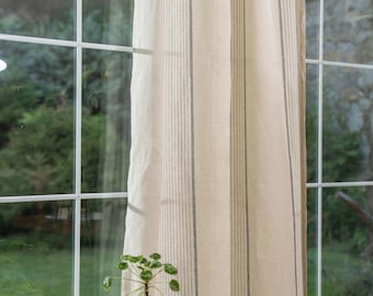 Puede incluir: Un par de cortinas de lino beige con finas rayas grises cuelgan frente a una ventana. Una pequeña planta en maceta se encuentra sobre un taburete negro delante de las cortinas.