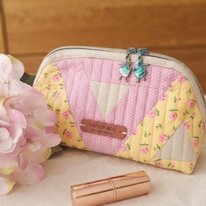 Könnte beinhalten: Ein gestepptes Stofftäschchen mit einem Patchwork-Design aus rosa Punkten, gelben Blumenmustern und beigen Dreiecken. Es verfügt über zwei schillernde, feenförmige Reißverschlussanhänger und ein braunes Lederetikett mit der Aufschrift HANDMADE Limited edition.