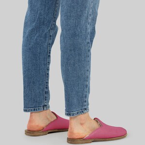 May include: A pair of fuchsia pink leather slippers with a rounded toe and a low, flat sole. The slippers are worn with blue denim jeans. The sole appears to be made of a light brown material.