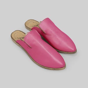 May include: A pair of bright pink leather slip-on shoes. The shoes have a rounded toe and a low heel. The soles are a light brown color. The shoes are displayed on a light gray background.
