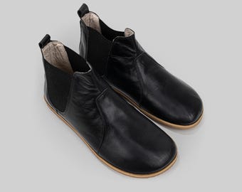 May include: A pair of black leather Chelsea boots. The boots feature a rounded toe, elastic side panels, and a pull tab at the back. The soles are light brown. The boots are shown against a light grey backdrop.