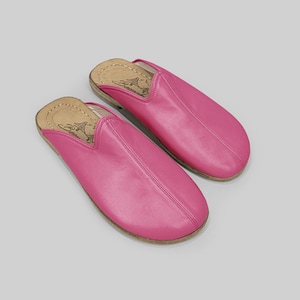 May include: A pair of bright pink leather slippers. The slippers have a rounded toe and a stitched seam down the center. The soles are a light brown color. The slippers are shown on a light gray background.