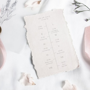 Könnte beinhalten: Eine weiße Papierkarte mit einem Zeitplan für die Ereignisse eines Hochzeitstages. Die Karte trägt das Datum "26.08.2017" und den Text "Der gro". Die Karte ist mit einem rosa Band und einem Wachssiegel verziert.