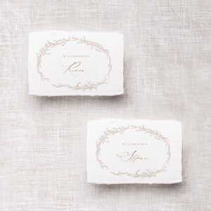May include: Two white rectangular place cards with a floral wreath design and the text "Willkommen" followed by a name in cursive. The top card says "Willkommen Rimen" and the bottom card says "Willkommen  Stefanie".