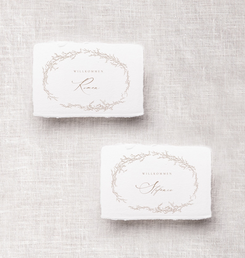May include: Two white rectangular place cards with a floral wreath design and the text "Willkommen" followed by a name in cursive. The top card says "Willkommen Rimen" and the bottom card says "Willkommen  Stefanie".