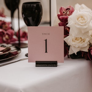 May include: A pink table number card with the word "Tisch" and the number "1" in black text. The card is sitting on a black stand and is surrounded by white roses and burgundy flowers.