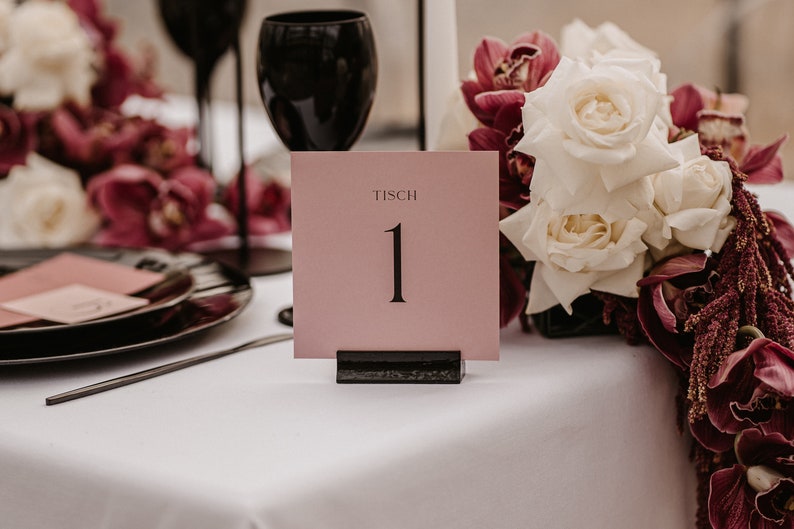 May include: A pink table number card with the word "Tisch" and the number "1" in black text. The card is sitting on a black stand and is surrounded by white roses and burgundy flowers.