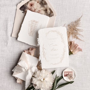 May include: Wedding invitation suite with a white background. The invitation features a delicate floral wreath with the names "Cassia & Richard" in calligraphy. The invitation is accompanied by a smaller card with a hand-drawn illustration of a building.