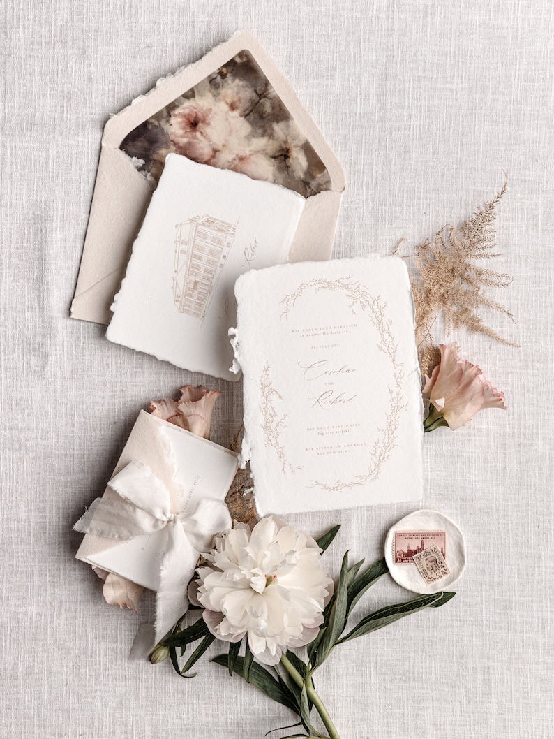 May include: Wedding invitation suite with a white background. The invitation features a delicate floral wreath with the names "Cassia & Richard" in calligraphy. The invitation is accompanied by a smaller card with a hand-drawn illustration of a building.