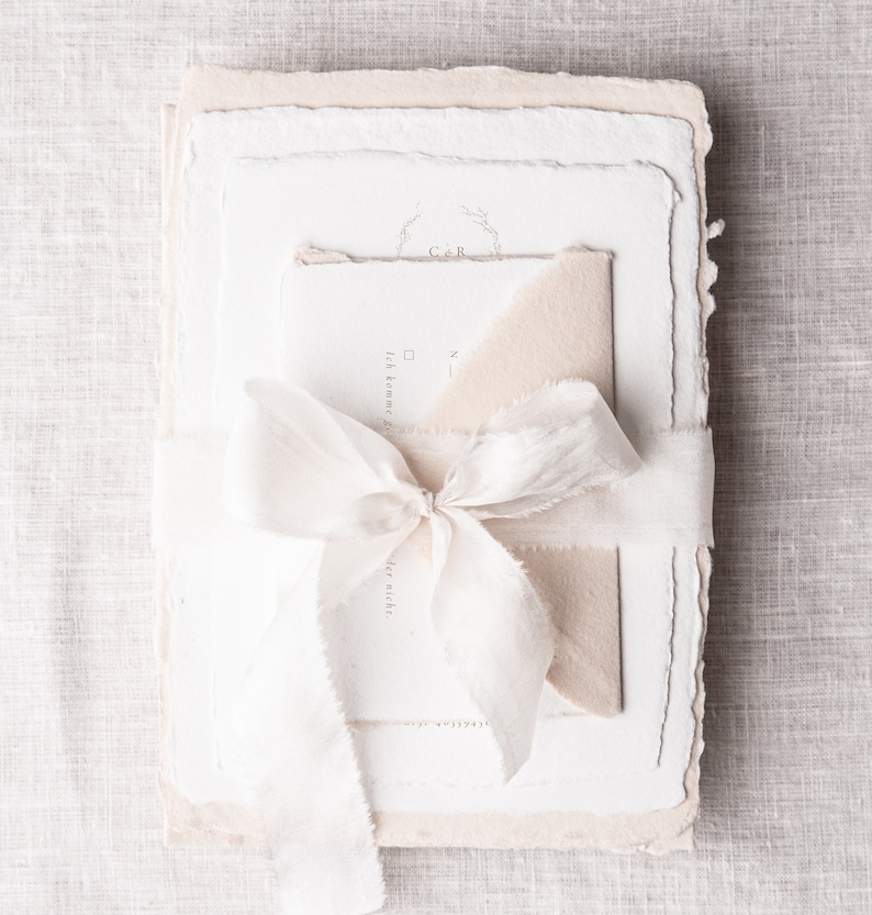 May include: A stack of white and cream paper stationery with a white ribbon tied around it. The top sheet has the initials "C&R" in a circle with a floral design. The text "Ich komme zu dir" is visible on the second sheet.
