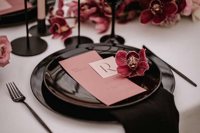 May include: A black plate with a pink menu card that says "Bon Appetit" and a single burgundy orchid on top. The plate is set on a white tablecloth with a black napkin.