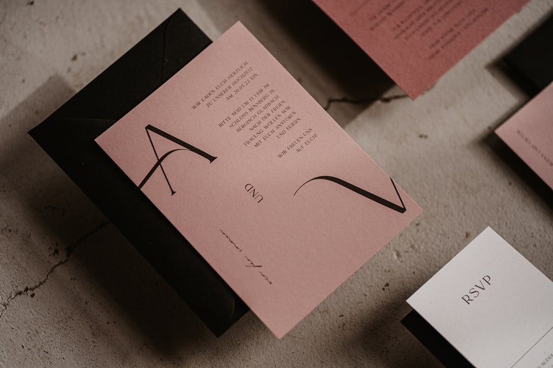 May include: A pink wedding invitation with black calligraphy lettering. The invitation includes the text "Wir laden Euch herzlich zu unserer Hochzeit am 20.05.22 in Bergisch Gladbach. Bitte seid um 11 Uhr im Bergischen Hof. Nach der Trauung wollen wir feiern mit Euch.  [Name] und [Name] Wir freuen uns auf Euch."