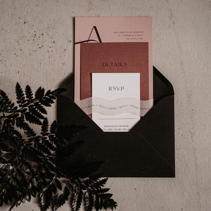 May include: A black envelope containing a pink and red invitation card. The invitation card features the text "RSVP" and "DETAILS". The envelope is resting on a light brown surface with a black fern branch to the left.