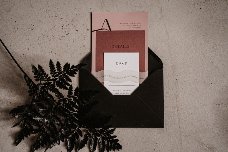 May include: A black envelope containing a pink and red invitation card. The invitation card features the text "RSVP" and "DETAILS". The envelope is resting on a light brown surface with a black fern branch to the left.