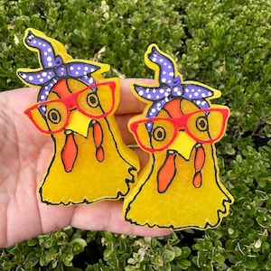 May include: Two yellow chicken-shaped ornaments with textured surfaces. Each chicken features a purple bandana with white polka dots, bright red glasses, and black outlines. The figures are crafted with a flat back and a vibrant yellow finish.