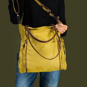 Pode incluir: Uma bolsa tote de lona amarela com al&ccedil;as e detalhes em couro marrom. A bolsa tem uma al&ccedil;a longa para carregar no ombro.