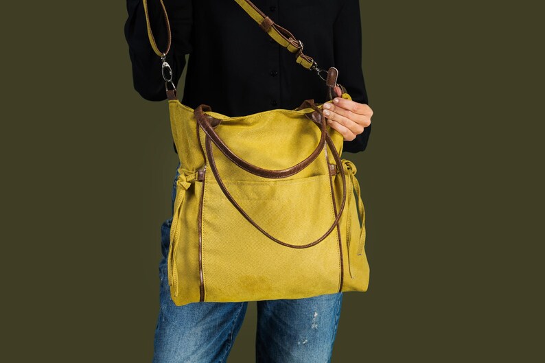 Pode incluir: Uma bolsa tote de lona amarela com al&ccedil;as e detalhes em couro marrom. A bolsa tem uma al&ccedil;a longa para carregar no ombro.