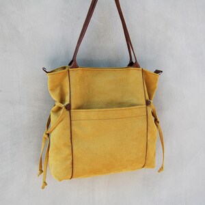 Pode incluir: Uma bolsa tote de lona amarela mostarda com al&ccedil;as de couro marrom e um grande bolso frontal. A bolsa tem fechos de cord&atilde;o nas laterais.