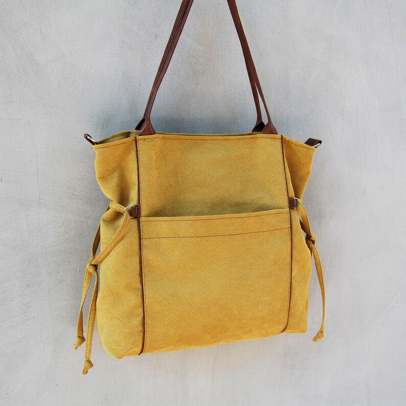 Pode incluir: Uma bolsa tote de lona amarela mostarda com al&ccedil;as de couro marrom e um grande bolso frontal. A bolsa tem fechos de cord&atilde;o nas laterais.