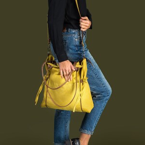 Pode incluir: Uma pessoa vestindo uma camisa preta e jeans azul est&aacute; carregando uma bolsa tote de lona amarela com detalhes em couro marrom e uma al&ccedil;a longa amarela.