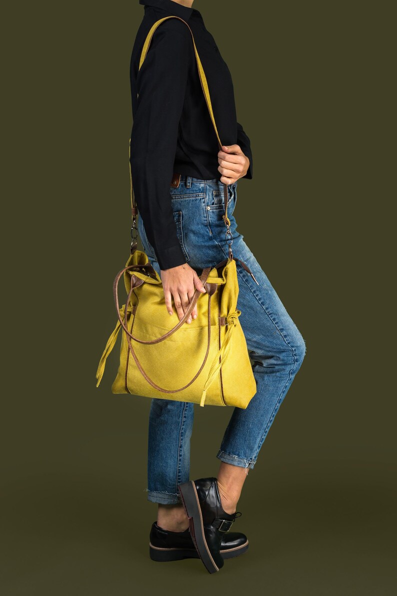 Pode incluir: Uma pessoa vestindo uma camisa preta e jeans azul est&aacute; carregando uma bolsa tote de lona amarela com detalhes em couro marrom e uma al&ccedil;a longa amarela.