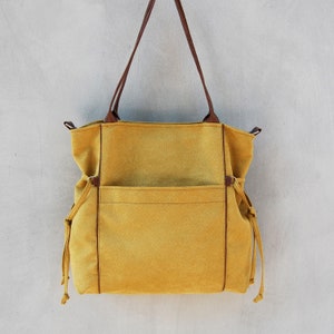 Pode incluir: Uma bolsa tote de lona amarela mostarda com al&ccedil;as de couro marrom e um grande bolso frontal. A bolsa tem fechos de cord&atilde;o nas laterais.