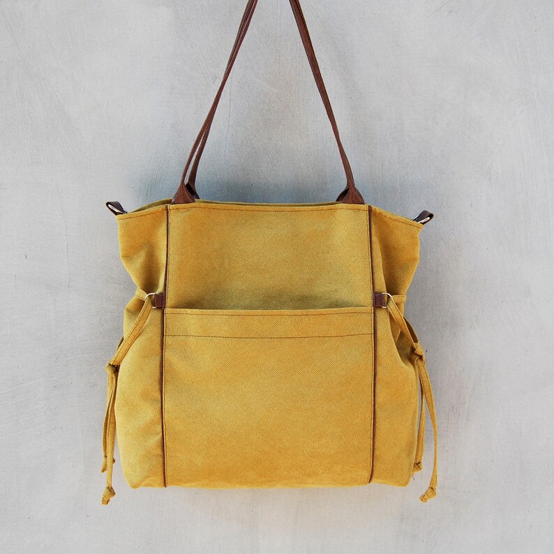 Pode incluir: Uma bolsa tote de lona amarela mostarda com al&ccedil;as de couro marrom e um grande bolso frontal. A bolsa tem fechos de cord&atilde;o nas laterais.