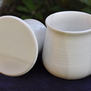 Könnte beinhalten: Eine weiße Keramik-Butterdose mit Deckel. Die Dose hat eine abgerundete Form und eine glatte, weiße Glasur. Der Deckel ist rund und hat einen leicht erhöhten Rand.