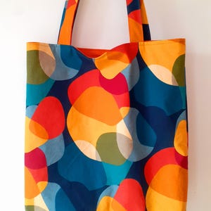 Könnte beinhalten: Eine farbenfrohe Tragetasche mit einem lebendigen, abstrakten Muster. Das Design zeigt sich überlappende Formen in Orange, Rot, Gelb, Blau und Grün auf einem dunkelblauen Hintergrund. Die Tasche hat passende orangefarbene Griffe und eine einfache, offene Oberseite.