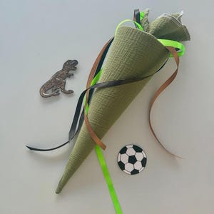 Può includere: Un sacco scolastico a forma di cono in tessuto verde con nastri colorati, un ritaglio di dinosauro e un adesivo a forma di pallone da calcio. La borsa è realizzata in un tessuto strutturato ed è riempita di tessuto verde. Sono attaccati nastri neri, marroni e verde neon.