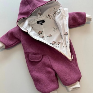 Peut inclure: Combinaison bébé prune avec capuche et fermeture éclair blanche. La doublure intérieure présente un motif de tortues et de feuilles sur fond blanc. La combinaison a des poignets blancs et une petite poche.