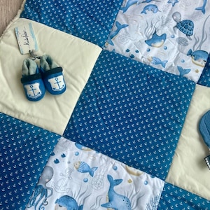 Könnte beinhalten: Ein Set aus Babydecke und Babyschuhen. Die Decke ist ein Patchwork-Design mit blauen Anker-Mustern und Illustrationen von Meeresbewohnern. Die blauen Babyschuhe haben Ankerembleme. Das Set ist für Babys konzipiert.