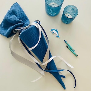 Op de afbeelding: Een blauwe, kegelvormige cadeauzak van stof, vastgebonden met witte en grijze linten, naast twee blauwe drinkglazen, een dolfijnsticker en twee blauwe kleurpotloden. De tas is gemaakt van een textuurstof.
