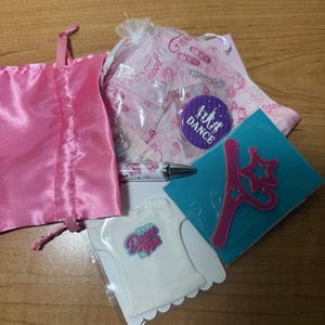May include: A collection of dance-themed items, including a pink satin bag, a white t-shirt with "Dance Top" text, a pen, a button with "Dance" text, and a blue card with a pink star-shaped wand.