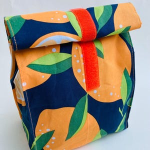 Puede incluir: Una bolsa de almuerzo reutilizable con un vibrante estampado de cítricos naranja y azul marino. La bolsa tiene un cierre enrollable asegurado por una correa de velcro naranja brillante. Hojas verdes y puntos blancos acentúan el diseño de la fruta naranja.