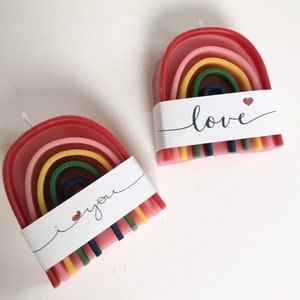 Könnte beinhalten: Zwei regenbogenförmige Kerzen mit einem weißen Band. Auf einem Band steht "i you" mit einem Herz, auf dem anderen "love" mit einem Herz. Die Kerzen haben Schichten aus rotem, rosa, gelbem, grünem und blauem Wachs. Die Kerzen stehen auf einer weißen Oberfläche.