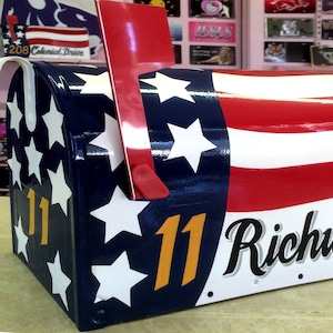 May include: A red, white, and blue mailbox with a flag design. The mailbox has a white background with red and blue stripes and white stars. The mailbox has the number "11" and the address "Richwood Place" written on it.