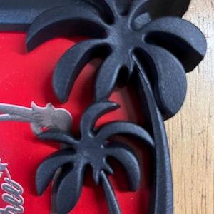 May include: Black picture frame with a tropical palm tree design. The frame has a matte finish and a red background. The palm trees are three-dimensional and add a decorative element to the frame.