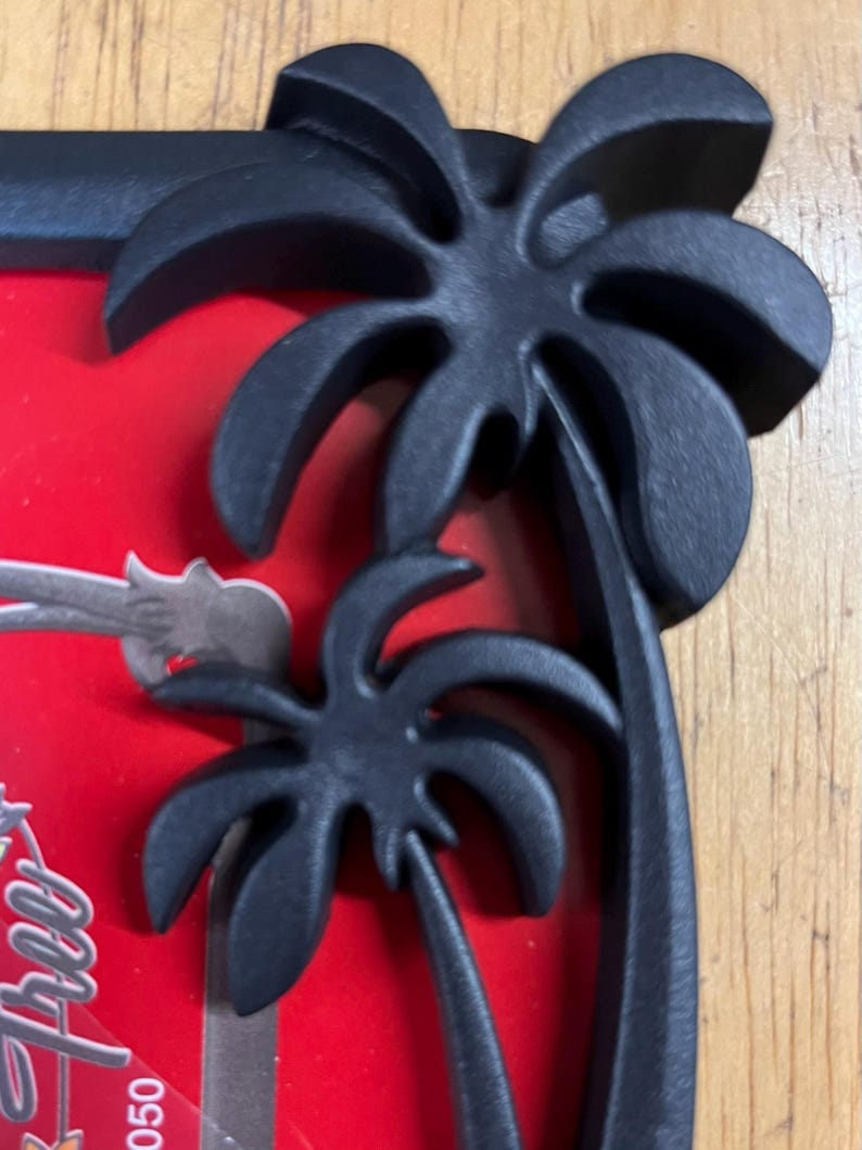 May include: Black picture frame with a tropical palm tree design. The frame has a matte finish and a red background. The palm trees are three-dimensional and add a decorative element to the frame.