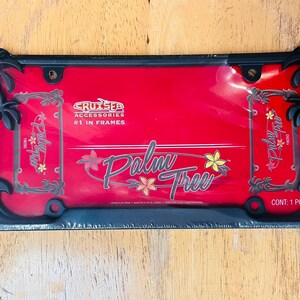 May include: Black license plate frame with a red background. The frame features palm tree cutouts at each corner and the words "Palm Tree" in a stylized font. The text "Cruiser Accessories #1 in Frames" is also visible.