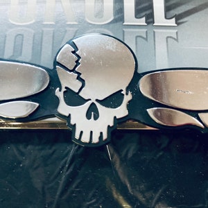 May include: A silver skull with a cracked design, attached to a black metal base with silver flames on either side.