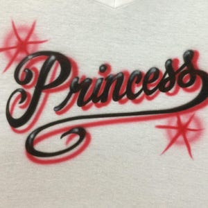May include: White v-neck t-shirt with the word "Princess" in black script with a red outline. Red starburst designs are on either side of the word. The shirt is made of a soft, textured material.