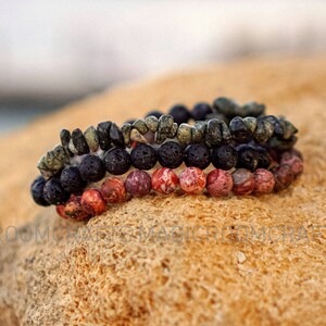 May include: Three stacked bracelets made of black lava rock beads, red and green jasper beads, and dark green and black speckled beads.