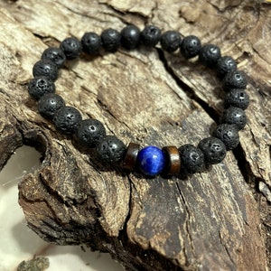 Può includere: Un braccialetto con perline di roccia vulcanica nera, una perla di occhio di tigre blu e distanziatori in legno.